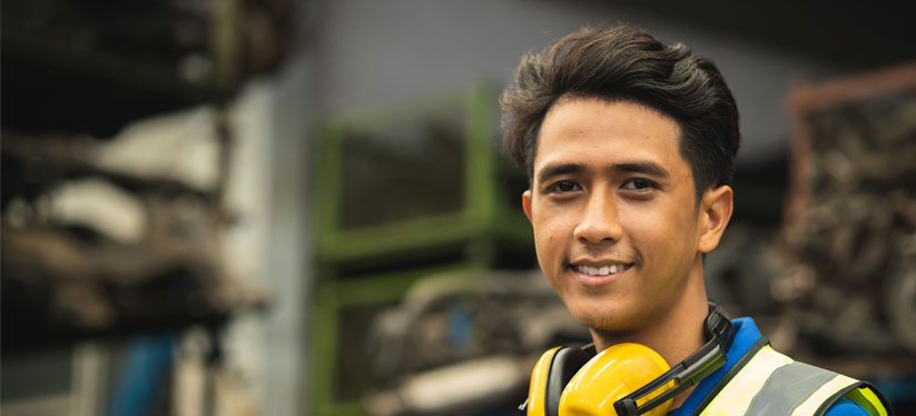 A smiling young man on a reflective vest with hearing protection earmuffs in his neck, looking to the left from the right side, over a blurred background